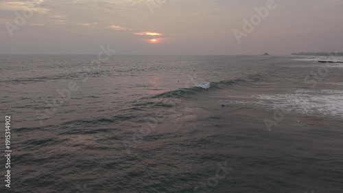 Surfers riding ocean waves during a golden sunset at Naomi Beach in Sri Lanka. Silhouettes of surfers against dramatic orange sky, tropical coastline, peaceful evening atmosphere, and authentic island
