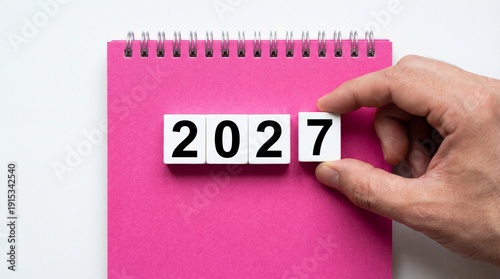 A hand placing the year 2027 on a pink notebook