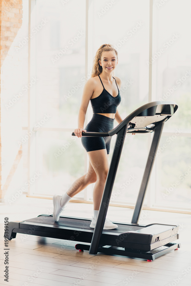 Fototapeta premium Young woman exercises on a treadmill in a bright gym with modern equipment and comfortable surroundings, enjoying her fitness routine