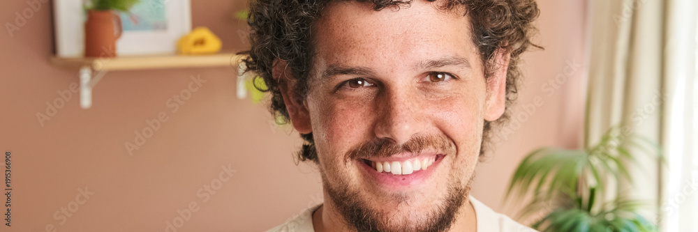 Obraz premium Close-up of young man with curly hair and beard looking at the camera with smile while standing in cozy living room. Panoramic