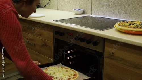 Young woman carefully placing a baking tray with homemade pizza into the oven. 