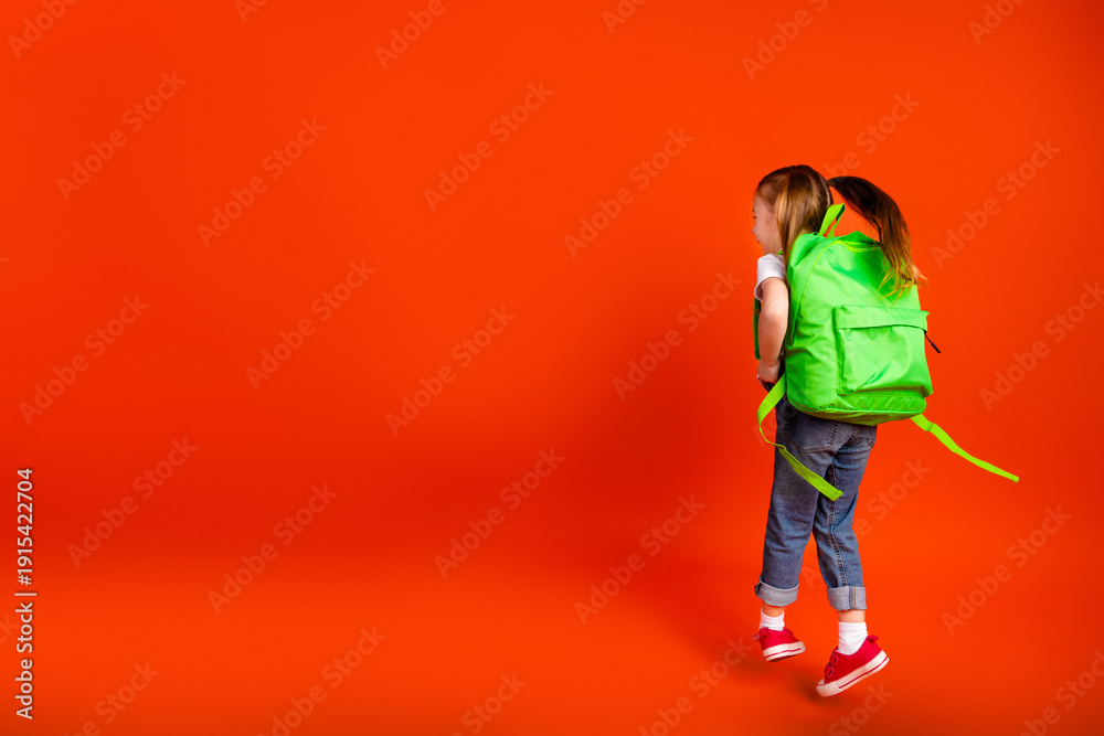 Naklejka premium Young girl with bright green backpack walking against bold orange background showcasing school fashion joy and family friendly vibes