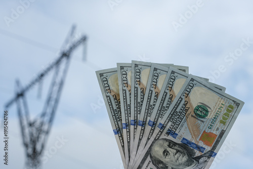 Close-up of one hundred US dollar bills against a blurred high voltage transmission tower. Electricity prices, energy market volatility, power infrastructure investment, and global energy transition.