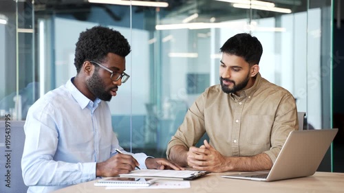 Sales manager sitting at office desk signing contract with client and shaking hands after successful deal. Multiethnic business partners confirming agreement, cooperation and trust in modern workspace