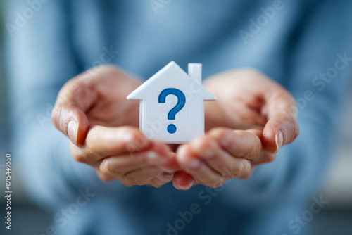 Person holding a small white house figure with a blue question mark symbolizing uncertainty or questions about homeownership or real estate decisions