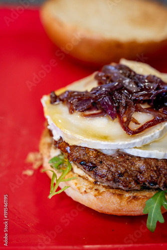 Fotografie Gros plan sur un burger gourmet ouvert garni d’un steak haché grillé, fromage fondant type brie, oignons caramélisés et roquette sur plan de travail rouge