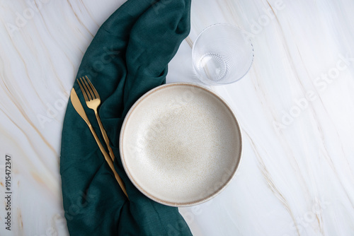 Beige ceramic plate with golden cutlery and drinking glass on light marble background with green fabric, horizontal minimalist composition with copy space. 
