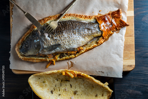 Whole baked fish wrapped in golden puff pastry served on wooden board with vegetables and cutlery, top view, closeup. Gourmet seafood dish concept