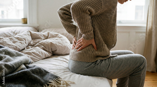 Woman sitting on bed while feeling intense lumbar pain and holding her lower back at home bedroom