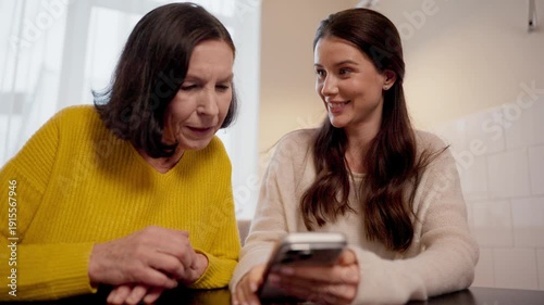 Pretty granddaughter showing photos on smartphone to experienced grandmother in bright kitchen at home. Sharing memories together while sitting close and smiling at screen with warm family connection.