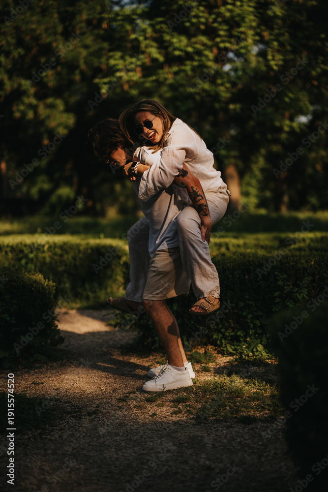 Fototapeta premium A couple shares a playful piggyback moment in a sunlit park. They smile and laugh as one carries the other along a garden path.