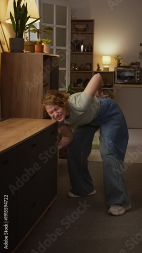 Full vertical shot of mature Caucasian woman moving and rearranging furniture in living room, grabbing cabinet, then collapsing with lower back injury, crying in pain and massaging lumbar area