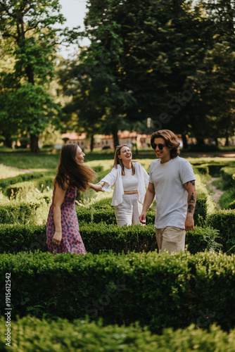 Fotografija Three friends laugh and converse as they walk through a neatly trimmed garden hedge maze, capturing a lively outdoor moment