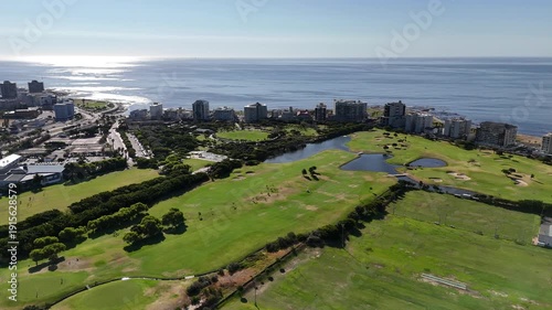 Drone flies west over sports fields, golf course, and park, toward the sea on a sunny day in Green Point, Cape Town, South Africa