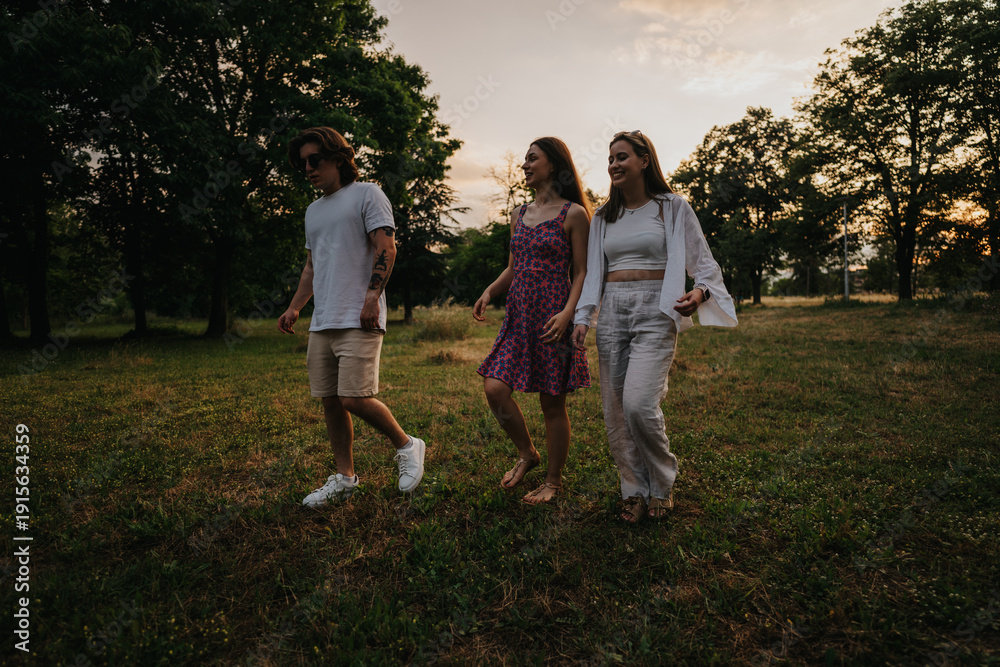 Fototapeta premium Three friends walk through a grassy park at sunset, smiling and relaxed. A warm outdoor scene suitable for lifestyle and friendship imagery.