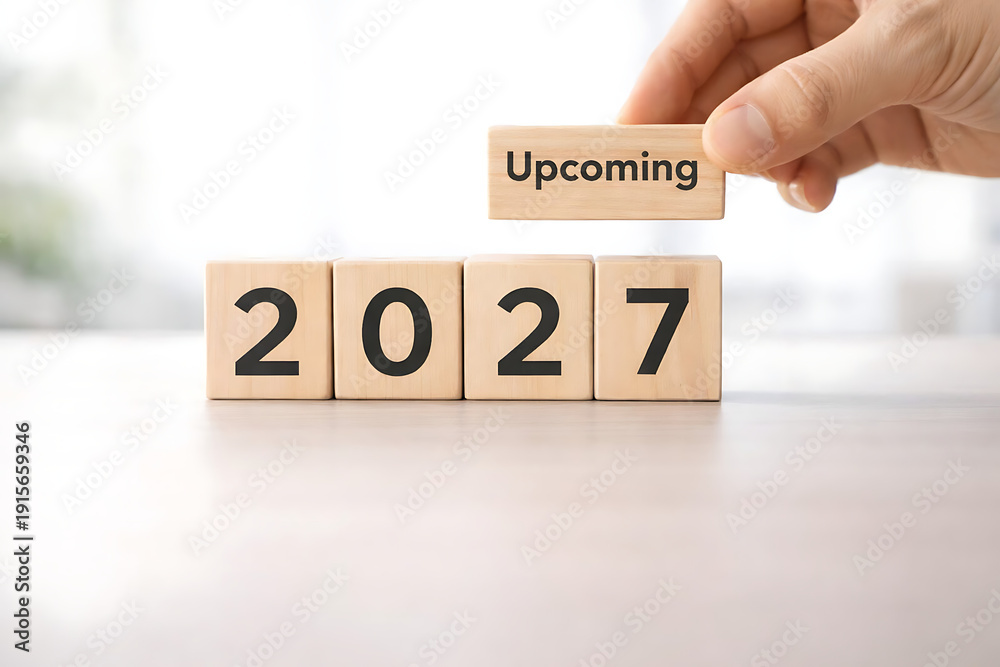 Naklejka premium Hand adding 'Upcoming' block to wooden blocks spelling '2027', representing future planning and New Year anticipation.