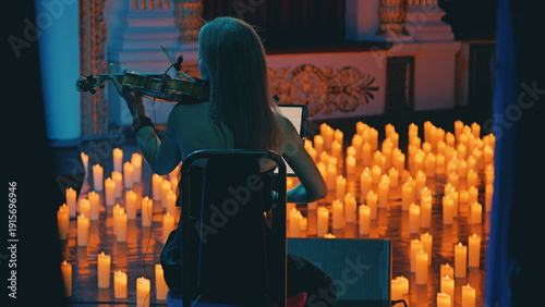 Over the shoulder view of a female musician playing violin in a romantic concert. Hundreds of candles illuminate the stage, creating a warm, magical atmosphere with contrasting cool blue tones