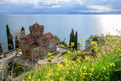 Photography Iconic Saint John Kaneo church on a rocky outcrop above Lake Ohrid, framed by cypress trees and vibrant spring wildflowers, under a partly cloudy natural light