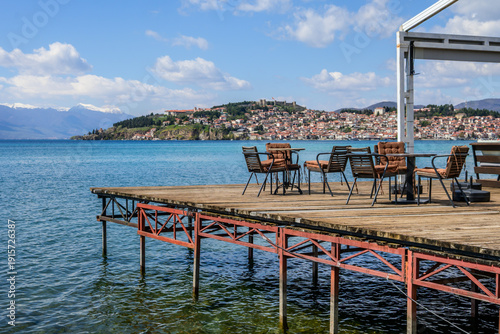 An inviting wooden cafe pier, complete with outdoor tables and chairs, offers stunning views of Ohrid's sprawling red-roofed city and distant snow-capped peaks across the calm blue lake.