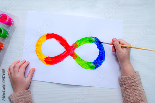 Top view of children’s hands painting an infinity symbol in rainbow colors. Creative activity supports child development, fine motor skills, imagination, represents autism awareness neurodiversity.