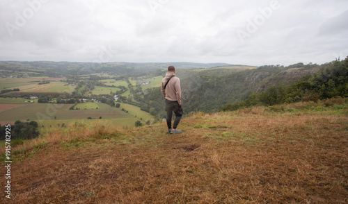 Male traveler standing at scenic overlook above a valley, wide countryside panorama in cloudy weather, freedom concept.