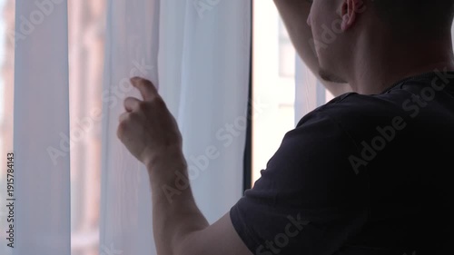 A man stands by the window, touching the sheer curtains.
Interior of a room.
White silk curtains in a cozy room.