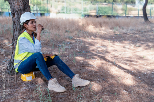 Woman worker with safety vest and hard hat resting under a tree