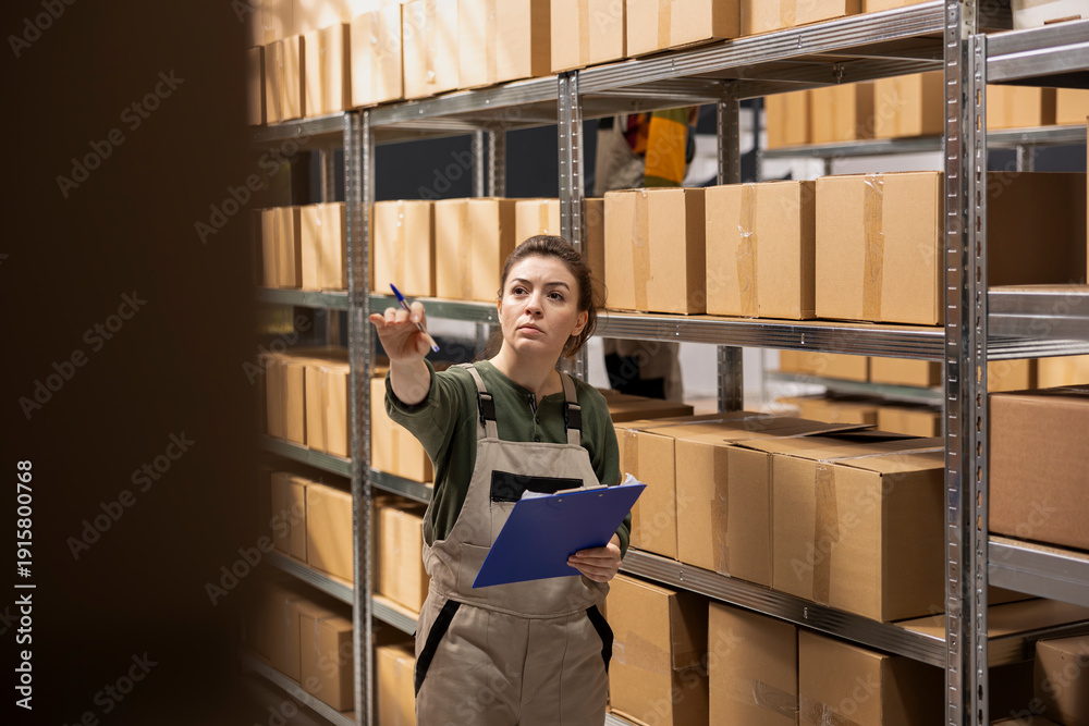 Fototapeta premium Woman staff member preparing goods for shipment in a small scale depot, handling e-commerce cargo and ensuring express delivery services. Checking parcels with inventory list clipboard.