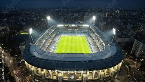 Aerial footage of a modern illuminated soccer stadium with green field at night, surrounded by a vast urban cityscape with numerous city lights