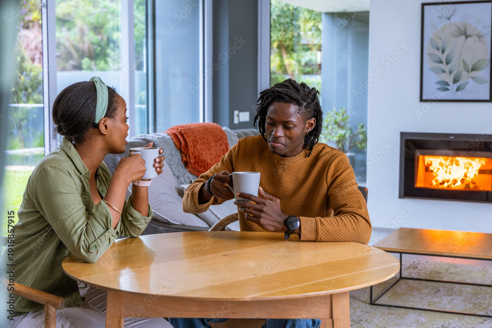 Fototapeta premium African American couple sitting at table holding mugs, chatting by lit fireplace and sliding doors