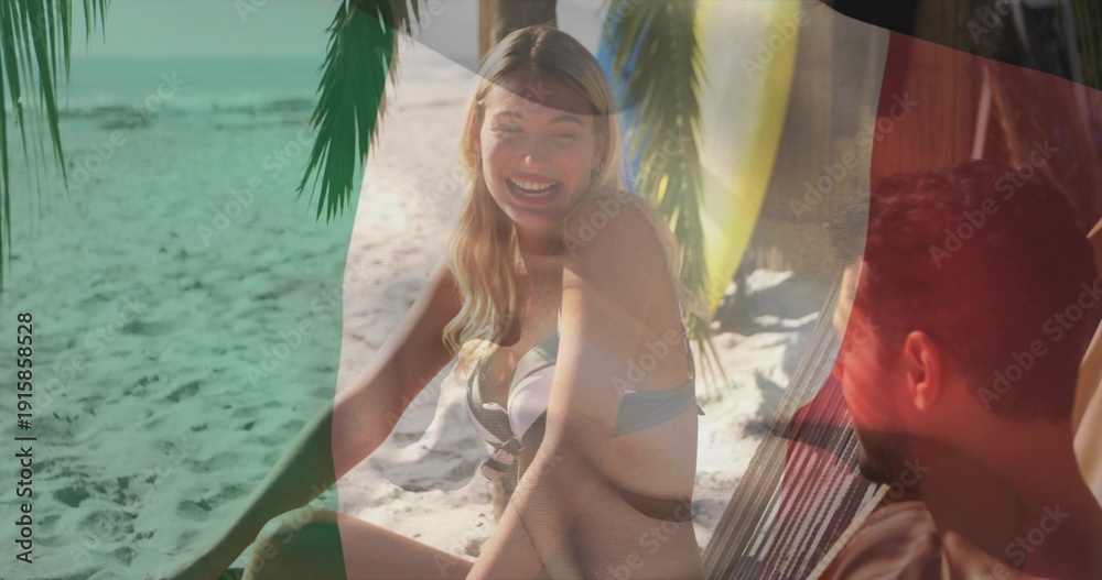 Fototapeta premium Laughing woman wearing bikini sitting on sand at tropical beach, with hammock and yellow surfboard