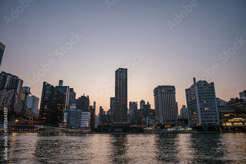 Wallpaper Mural Tall buildings of Manhattan rise along the East River during dusk, with city lights beginning to glow. The calm water reflects the skyscrapers and the fading evening sky. Torontodigital.ca