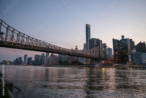 The Queensboro Bridge spans across the East River as Manhattan's illuminated skyline reflects in the calm water. The scene captures the peaceful beauty of the city during the last light of day.