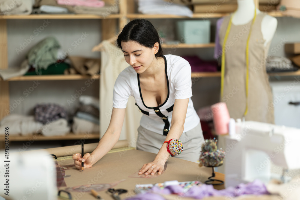 Fototapeta premium Dedicated young female dress designer working in creative fashion studio, developing new patterns, drawing and adjusting layout for future garment on paper using curve ruler