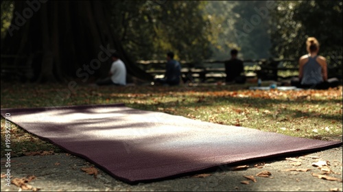 A textured yoga mat lies on the ground outdoors, with people meditating or practicing mindfulness in the background among trees and fallen leaves.