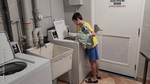 Young man removes wet clean laundry from a washing machine and transfers it into a dryer. The action shows routine household work, laundry care, and use of home appliances.