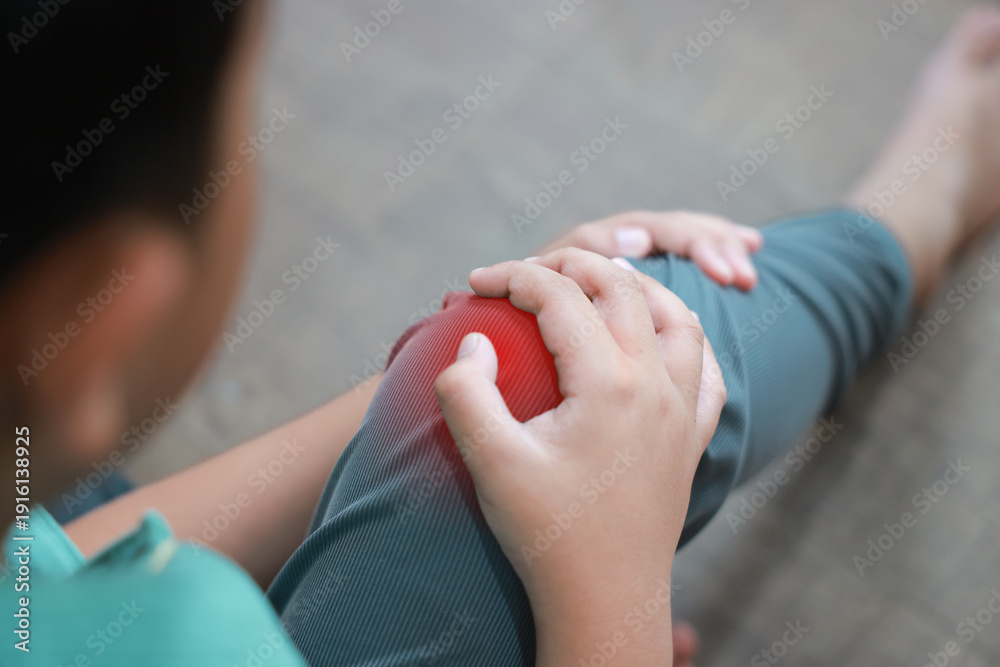 Naklejka premium Little boy holding red inflamed knee with hands suffering from joint pain or accidental injury at home. Concept of pediatric healthcare, bone problems in children, and orthopedic medical treatment.