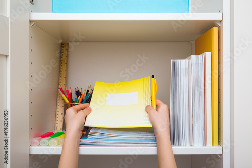 Little child hands holding copybook and pen. Different school accessories on white wardrobe shelf. Closeup. Making neatness. Point of view shot. Front view.