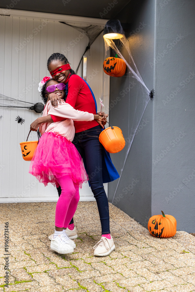 Naklejka premium African American mother and daughter hugging and posing on porch, holding orange pumpkin buckets
