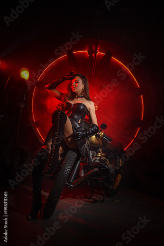 Stylish beautiful young woman in black leather corset and boots posing on vintage motorcycle on old garage dark interior background.