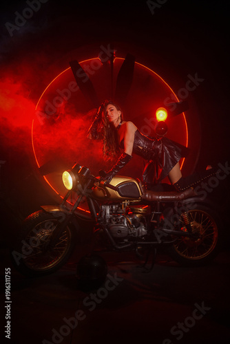 Stylish beautiful young woman in black leather corset and boots posing on vintage motorcycle on old garage dark interior background.