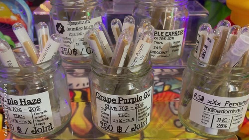 Close-up of labeled cannabis pre-rolled joints displayed in glass jars at a dispensary, showcasing different strains, THC levels, and sativa–indica profiles