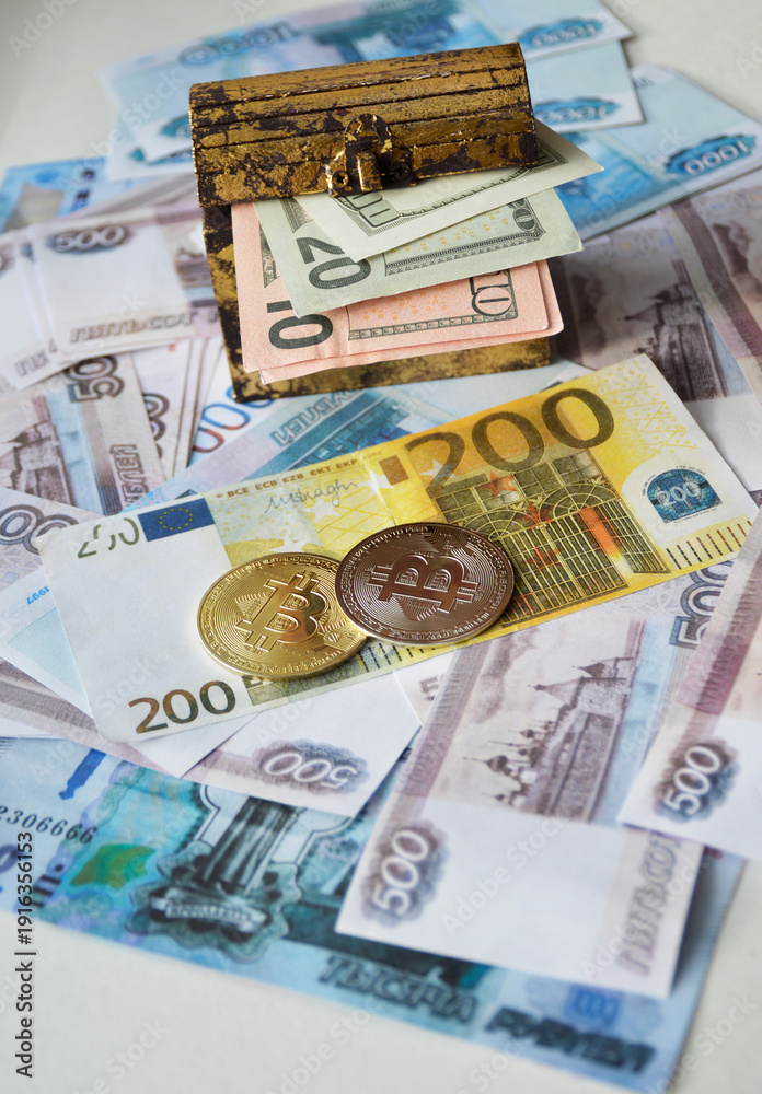 Obraz premium Two Bitcoin coins, a chest of dollars, and a Euro banknote against a background of Russian rubles. Concept of the transition to cashless payments and digital money. Trading. Cryptocurrency mining.