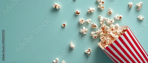 Red and white striped container with popcorn spilling onto a light teal surface; scattered popped kernels create a casual, fun impression. Perfect for themes related to cinema snacks