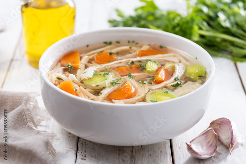 Homemade chicken soup in white bowl with carrots and celery on white wooden table
