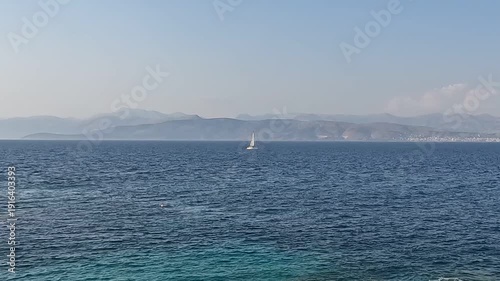 Single white sailboat navigating deep blue sea with hazy mountain backdrop