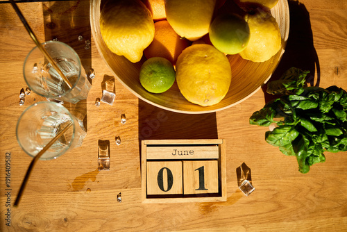 Fotografie A wooden bowl filled with colorful citrus fruits sits alongside two glasses with ice and fresh mint leaves, marking the start of June with a warm invitation to refreshment
