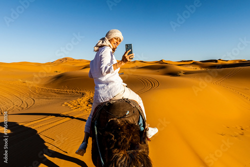 A woman in white clothing and headscarf rides a camel, taking a photo with her phone. Sand dunes stretch to the horizon under a clear blue sky. Mezouga,Sahara desert,Morocco