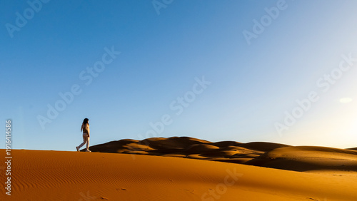 A woman walks along the crest of a large sand dune under a clear blue sky. Mezouga,Sahara desert,Morocco