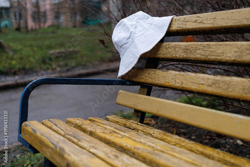 A white hat is placed on a yellow-painted wooden bench in a park. Green grass surrounds the area